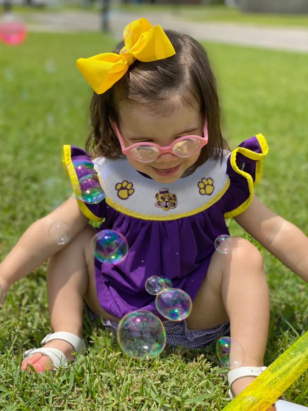Toddler girl with brown hair & bright yellow bow sitting looking at bubbles wearing purple bishop top with tiger smock on top