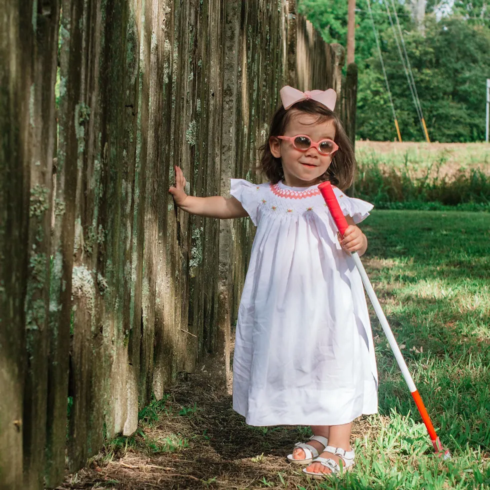 Toddler standing with right hand against fence looking to her left. She is holding a pink cane & wearing a pink bow & glasses