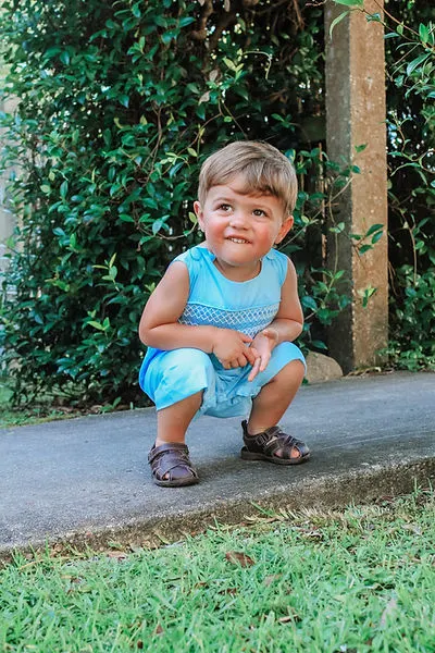Toddler boy crouches down and smiles wearing bright blue short-all