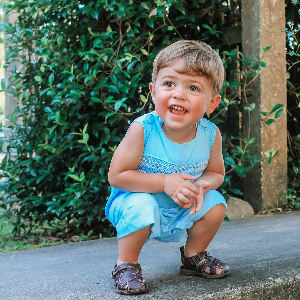 Toddler boy squatting and smiling wearing a baby blue shortall