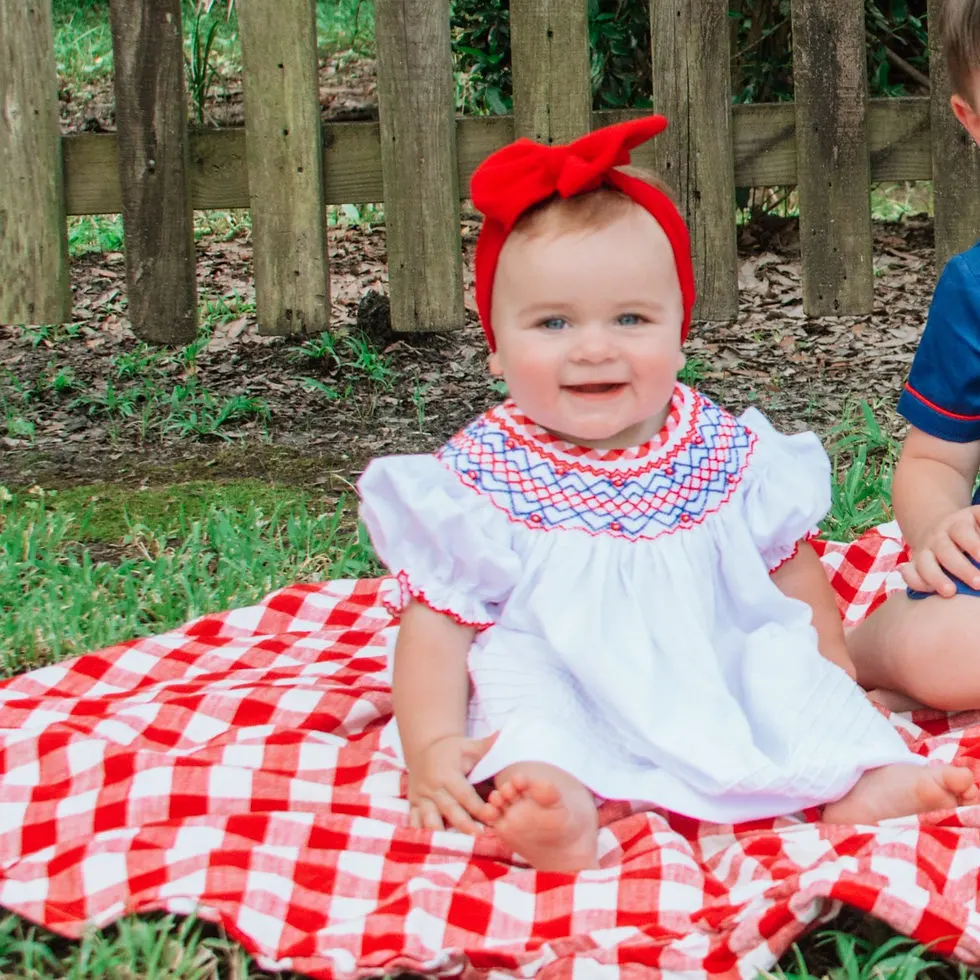 Baby girl wearing red headwrap & white dress with red & blue smocking across the chest sitting on a red & white plaid blanket