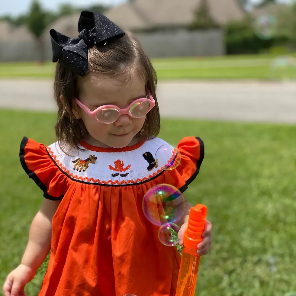 Toddler girl with brown hair & black sparkly bow with a bright orange bishop shirt & cowboy smock design across the top.