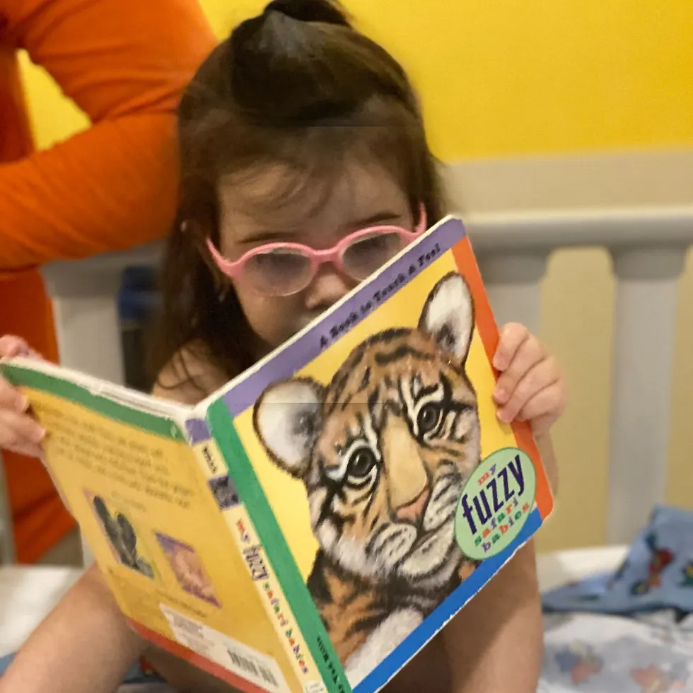 Toddler girl wearing pink glasses sits in hospital bed reading book titled My Fuzzy Safari Babies