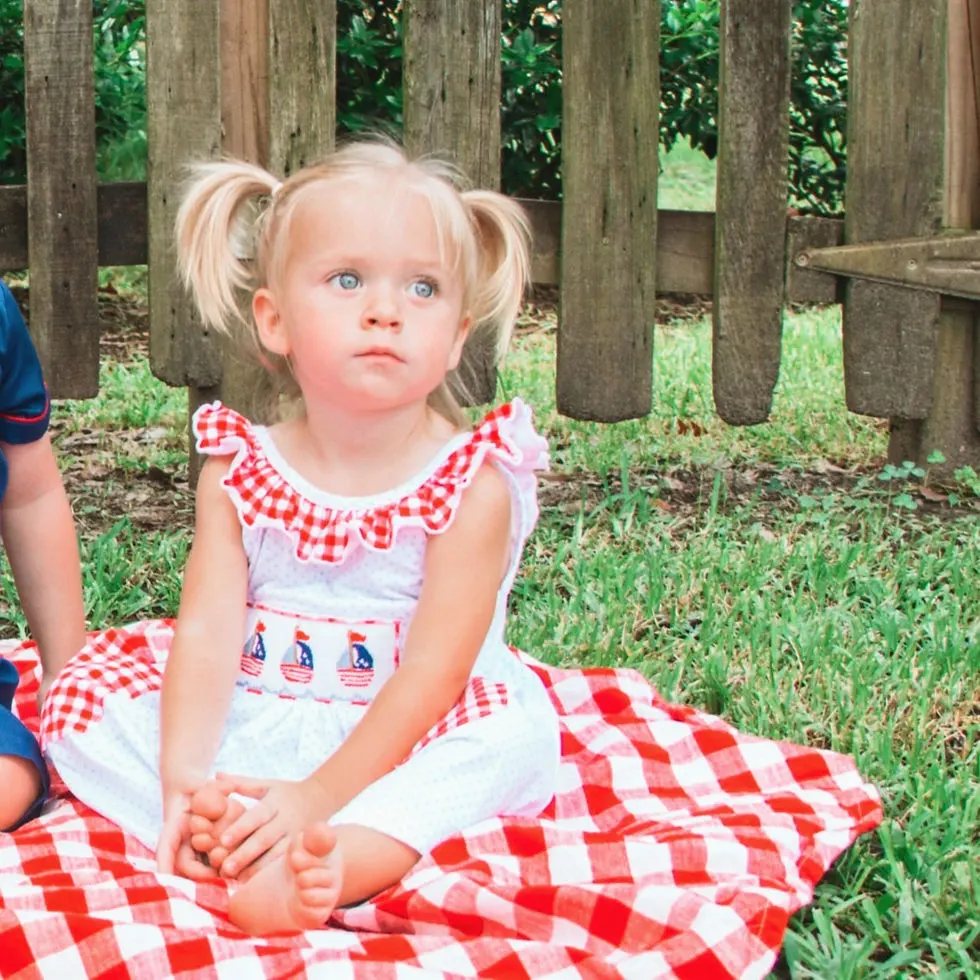Blonde haired girl wearing pigtails & a white dress with red & a white plaid ruffle across the top & sailboats smock on chest