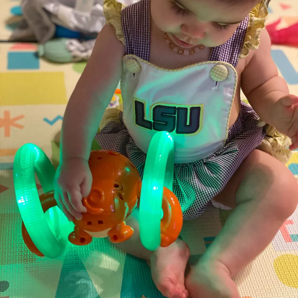 Infant girl wearing LSU bubble plays with monkey toy that rolls. The wheels are lit up green.