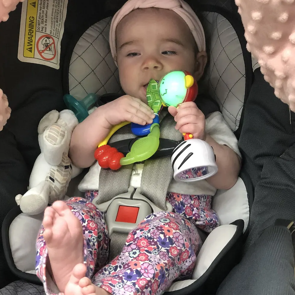 Infant girl in carseat holds teething toy in one hand and a rattle in the other that's lit up green