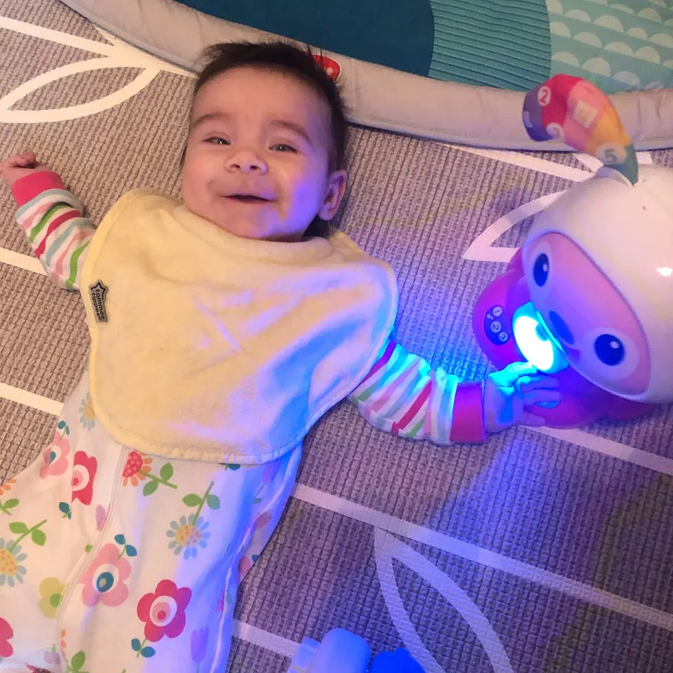 Infant girl lays on floor smiling and looking at camera. She's laying next to a toy with a bright circle in the middle.