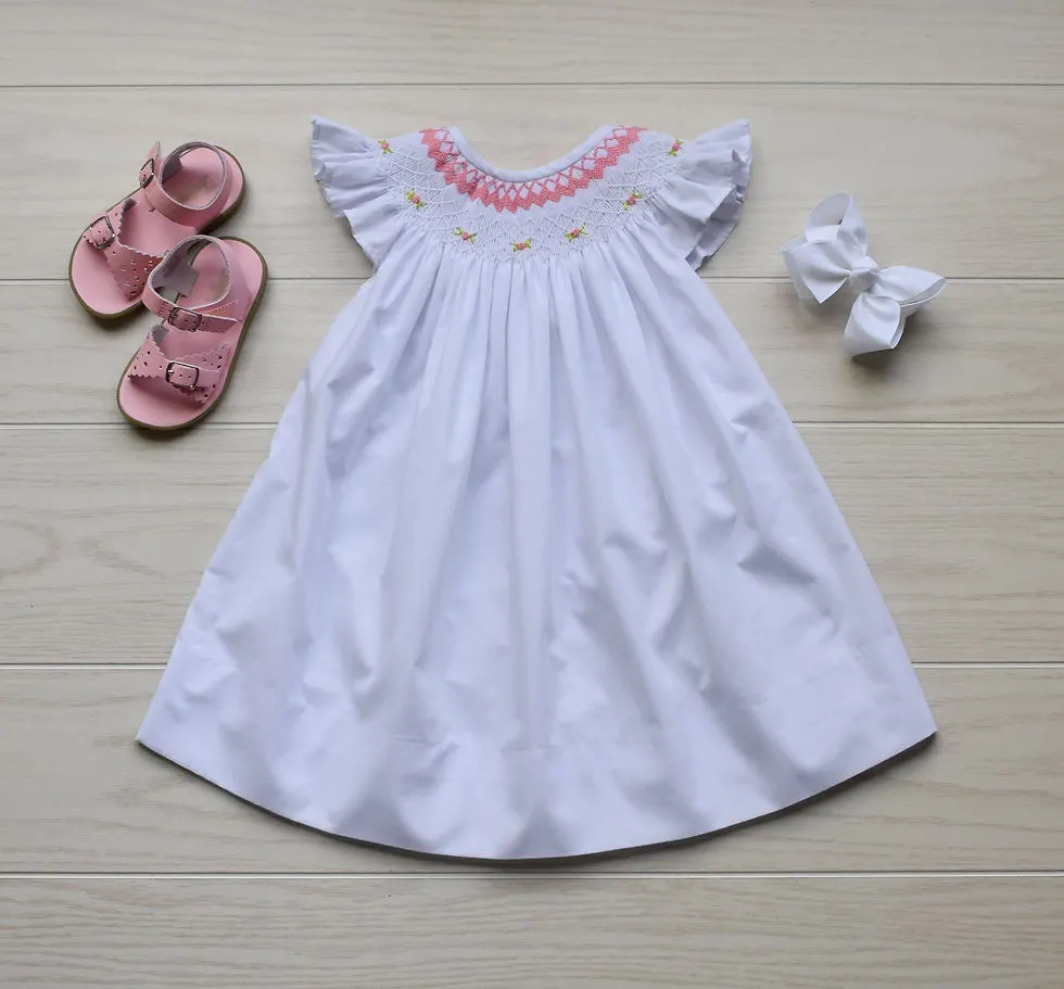 White dress with angled sleeves & pink & white smocking at the top. Pink shoes & a white bow lay on either side of the dress.