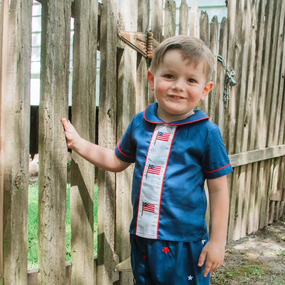 Young boy standing at fence smiling wearing a navy collared shirt with a white stripe down the middle with 3 American flags
